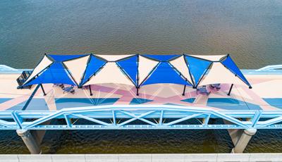 Blue and tan multi-panel shade structures covering tables and chairs on a bridge overlooking the Arkansas river. 