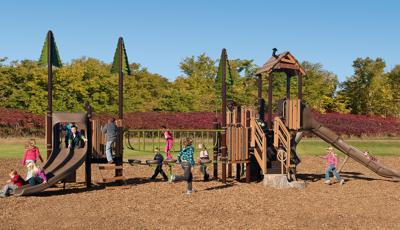 Kids playing on PlayBooster playground at Crimson Park. Littles ones line up for double slide, while two little girls ride toward the bottom. A girl runs across the play structure toward the stairs on the façade.