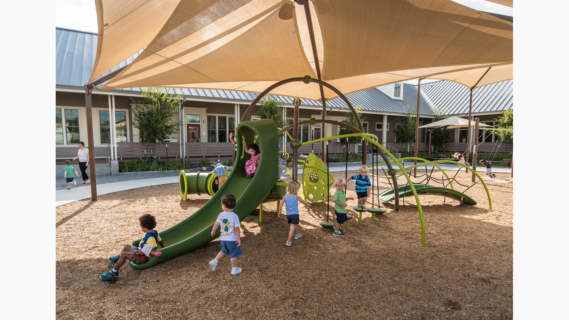 Toddler age children play on a small playground with climbers slides and tunnels all underneath a large tan shade system.