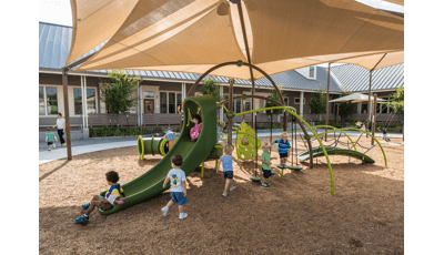 Toddler age children play on a small playground with climbers slides and tunnels all underneath a large tan shade system.