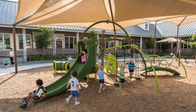 Toddler age children play on a small playground with climbers slides and tunnels all underneath a large tan shade system.