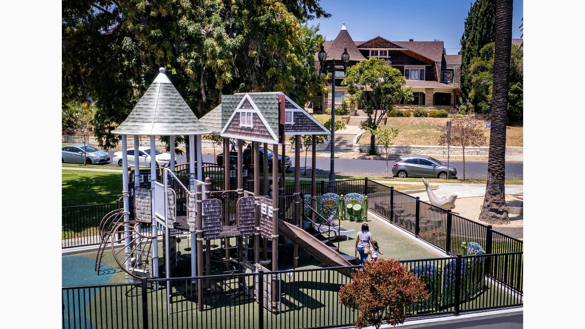 A fenced in play area with a custom designed house façade play structure set in a neighborhood setting.