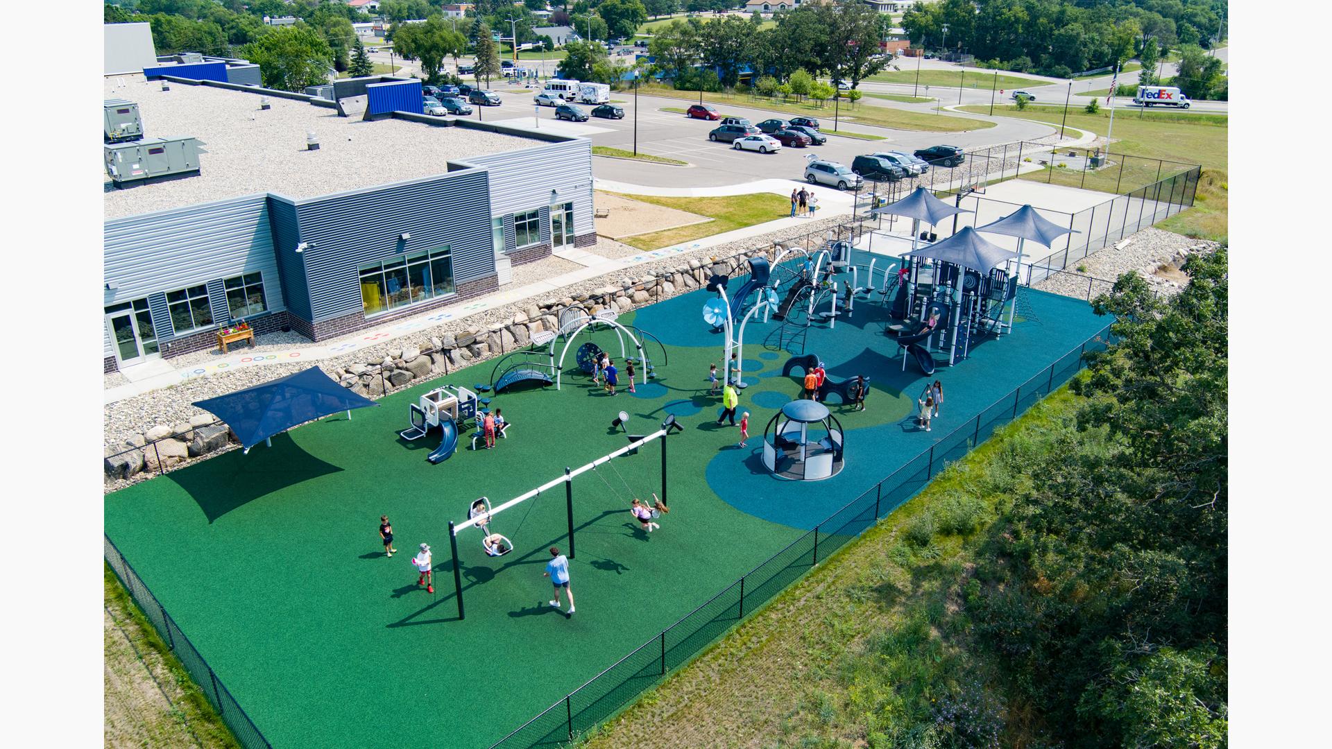 Elevated view of children playing on multiple play structures at an outdoor fenced in play area need to a community building.