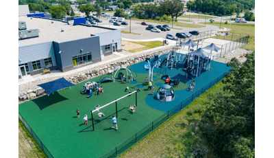 Elevated view of children playing on multiple play structures at an outdoor fenced in play area need to a community building.