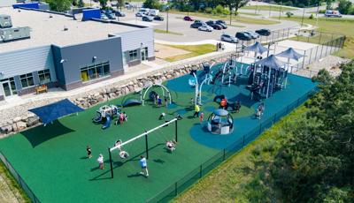 Elevated view of children playing on multiple play structures at an outdoor fenced in play area need to a community building.