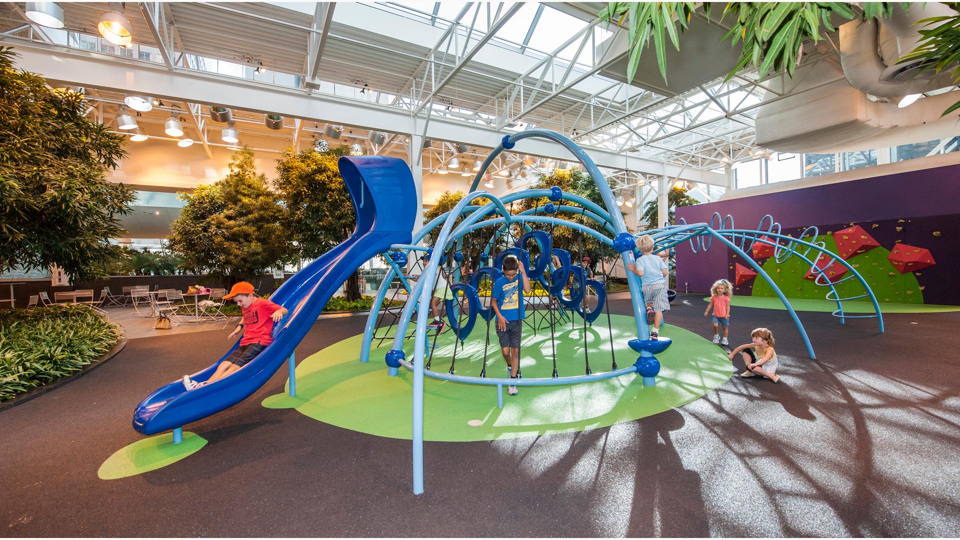 Indoor playground with rubber surfacing  and kid going down blue slide. Smaller kids playing on the floor while 2 kids play in net structure. Trees and greenery in background  with small place to eat. 