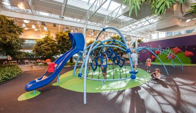 Indoor playground with rubber surfacing  and kid going down blue slide. Smaller kids playing on the floor while 2 kids play in net structure. Trees and greenery in background  with small place to eat. 