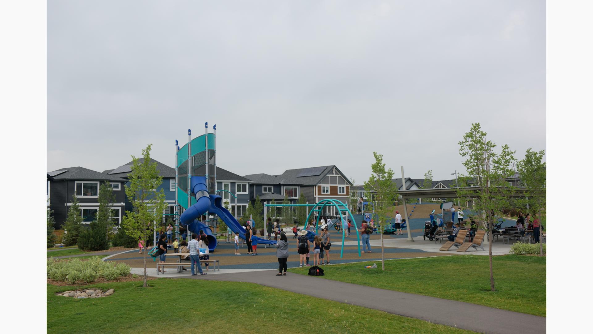 Families fill a play area of a neighborhood park with a main tower play structure and surrounding swing sets and multi person seesaw. 