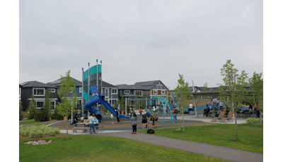 Families fill a play area of a neighborhood park with a main tower play structure and surrounding swing sets and multi person seesaw. 
