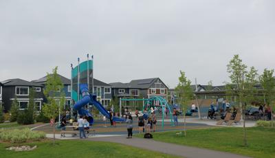 Families fill a play area of a neighborhood park with a main tower play structure and surrounding swing sets and multi person seesaw. 