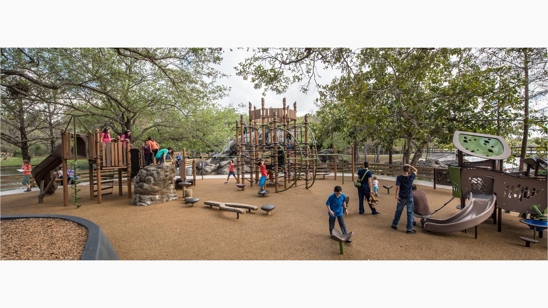 Families enjoy this nature-inspired playground. A father helps his toddler down a PlayShaper slide. A group of older kids play on the Geoplex net structure.