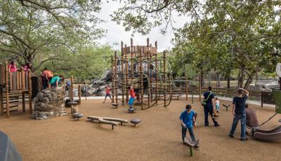 Families enjoy this nature-inspired playground. A father helps his toddler down a PlayShaper slide. A group of older kids play on the Geoplex net structure.