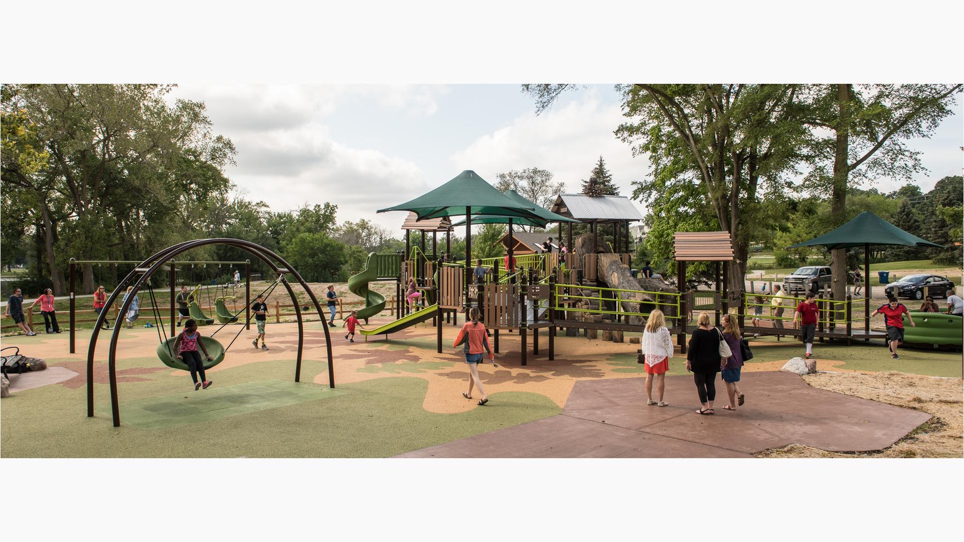This naturally-themed playground is full of children of all abilities accessing this playground's amenities. A girl sits on the Oodle swing. Parents enjoy the Sway fun and kids use the different ramps to get from one place to another.