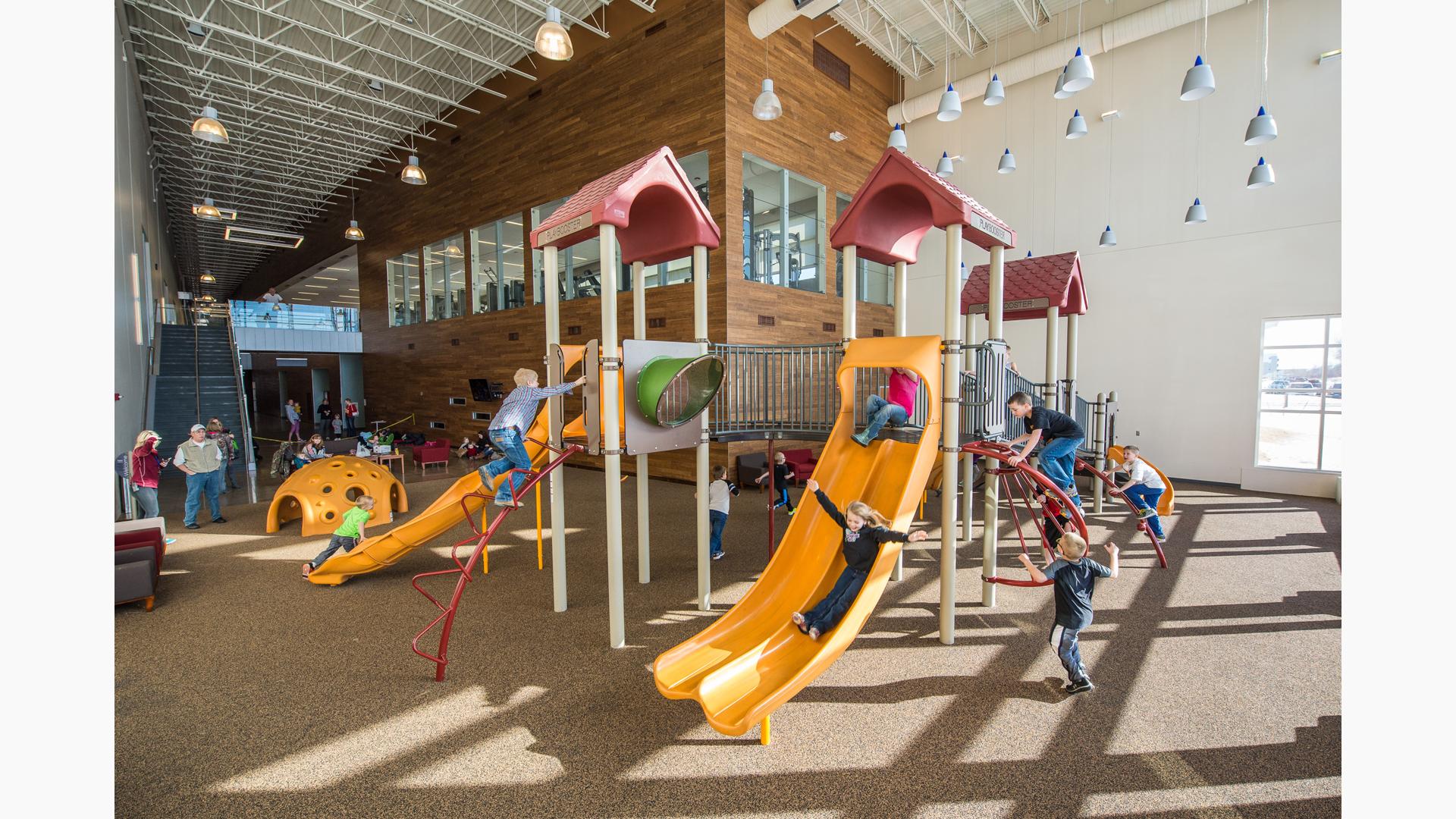 Girl in black sweater rides down slide, as a girl in a pink shirt sits at the top of the slide. Boys climb the Sunbeam climber, Croque climber, and lollipop climber. On the ground floor, children run around the play structure.