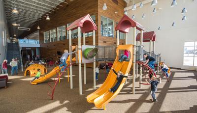 Girl in black sweater rides down slide, as a girl in a pink shirt sits at the top of the slide. Boys climb the Sunbeam climber, Croque climber, and lollipop climber. On the ground floor, children run around the play structure.