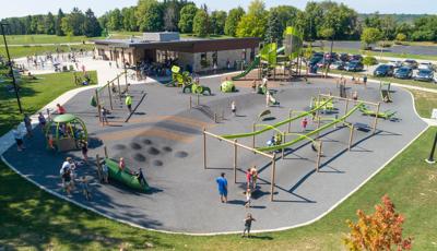 Families play at a large play area with accessible safety surfacing. Children play on ziplines, manual carousels, swing sets, and two play structures for older children and younger children. A brick building sits in the background with a small splash pad next to it.