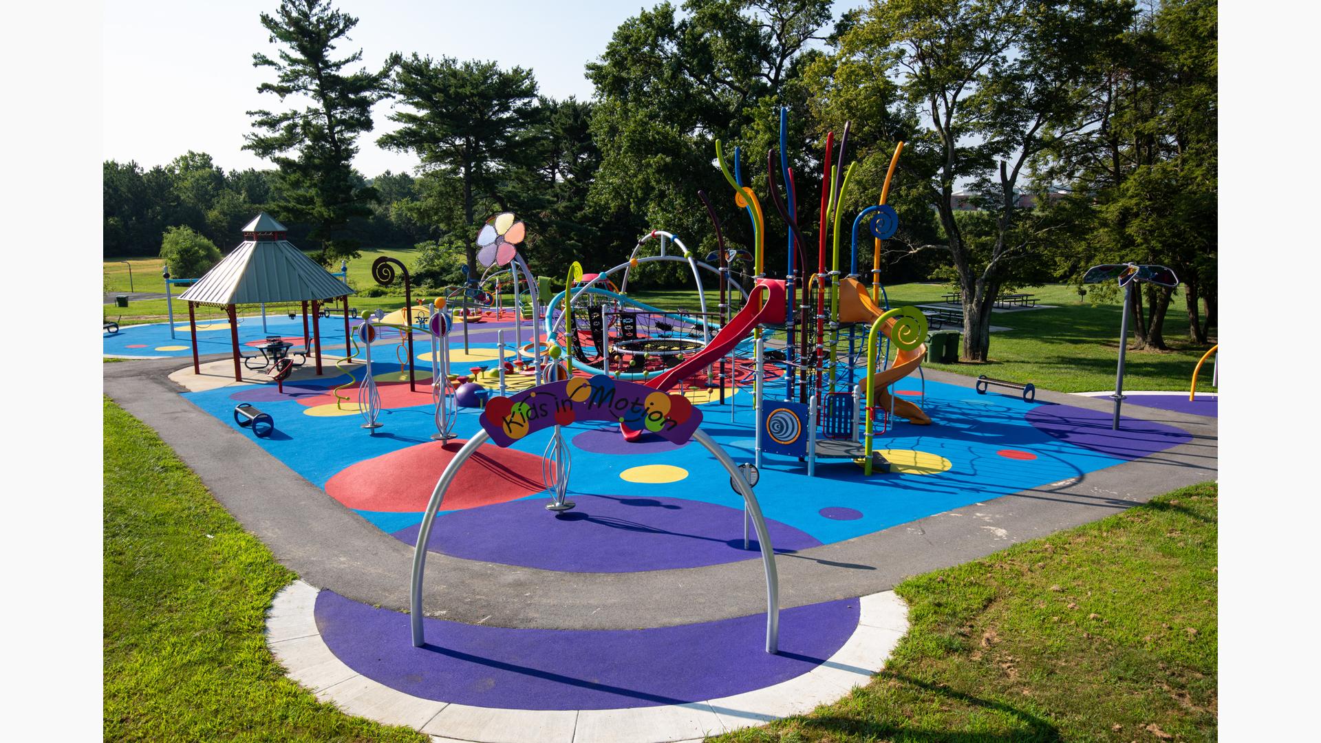 A rainbow color themed play area with play structures for younger and older children. Inclusive safety surfacing is designed in varying sized circles of multiple colors.