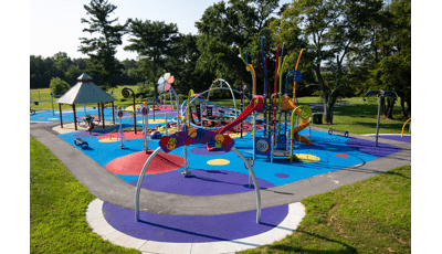 A rainbow color themed play area with play structures for younger and older children. Inclusive safety surfacing is designed in varying sized circles of multiple colors.