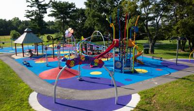 A rainbow color themed play area with play structures for younger and older children. Inclusive safety surfacing is designed in varying sized circles of multiple colors.