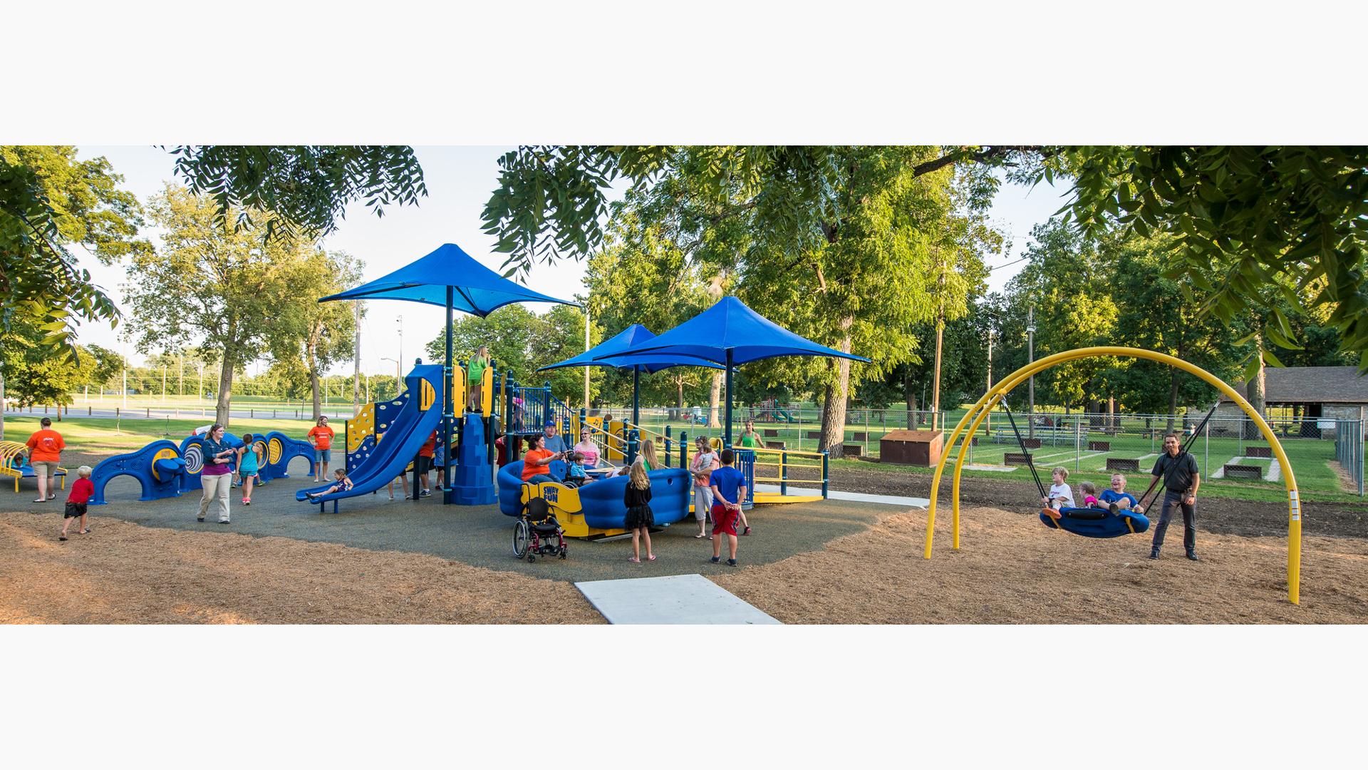 A dad pushes kids on a Oodle swing as ther families play on the playground.