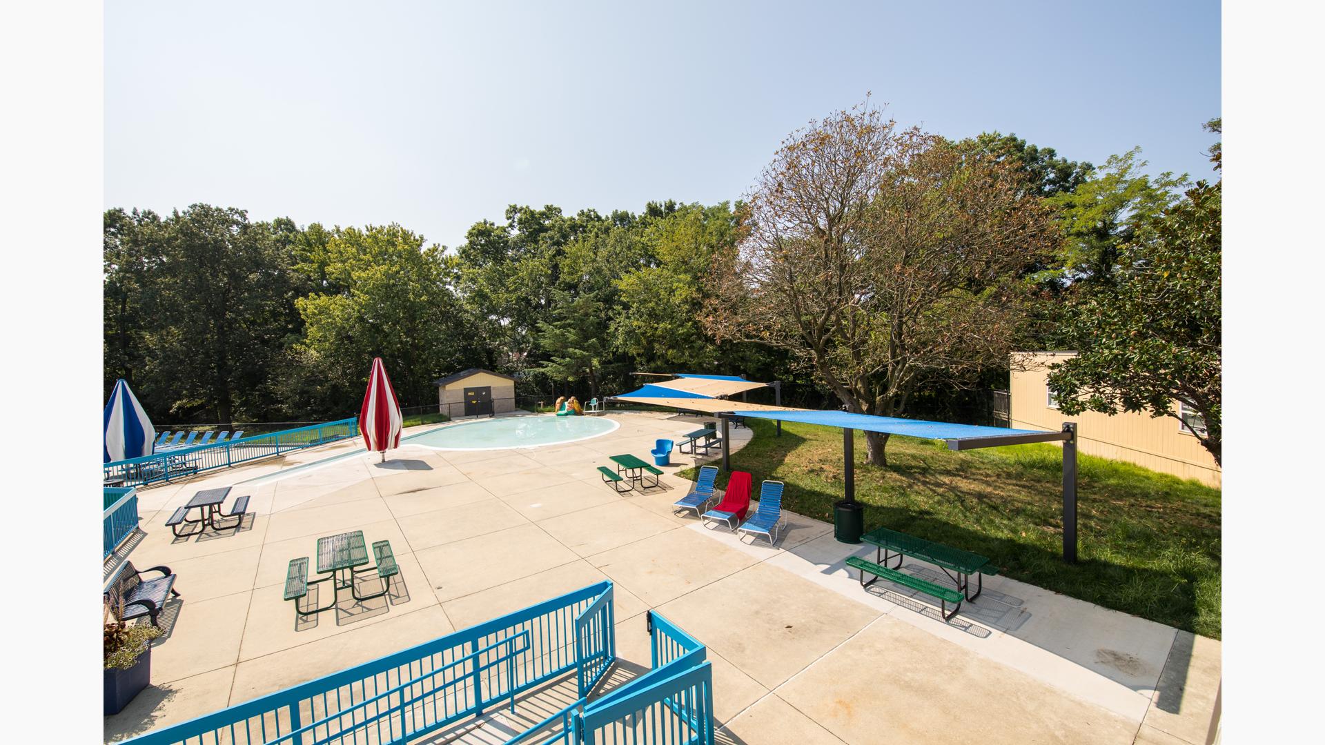 Swimming pool area with picnic tables, lounge chairs covered by tan and blue canopy shade structures. 