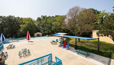 Swimming pool area with picnic tables, lounge chairs covered by tan and blue canopy shade structures. 