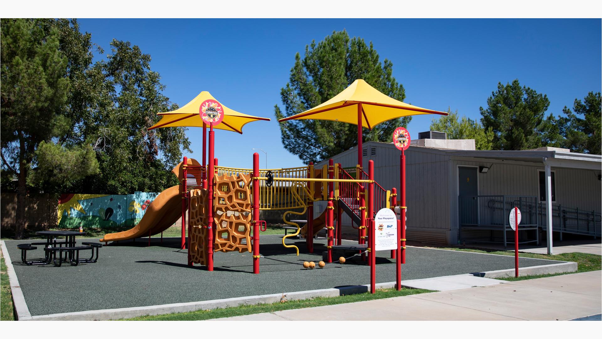 A play structure colored in red, yellows, and orange with a variety of play activities sit next to a building. Two bright yellow shades sit at the top of two of the playground posts. 