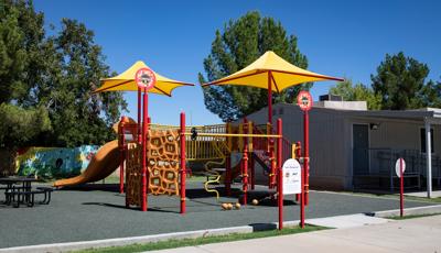 A play structure colored in red, yellows, and orange with a variety of play activities sit next to a building. Two bright yellow shades sit at the top of two of the playground posts. 