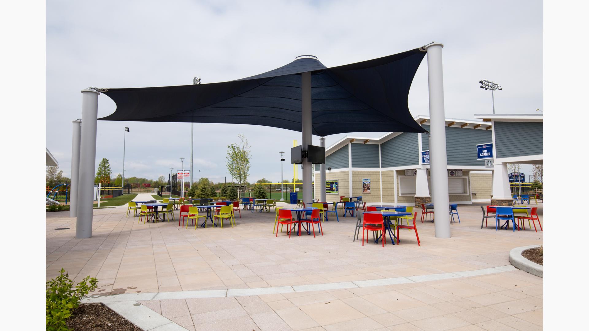 Tables and chairs of primary colors sit under a large navy blue custom shade structure with TV monitors on the center posts in the concession area of a sports center. 