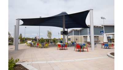 Tables and chairs of primary colors sit under a large navy blue custom shade structure with TV monitors on the center posts in the concession area of a sports center. 