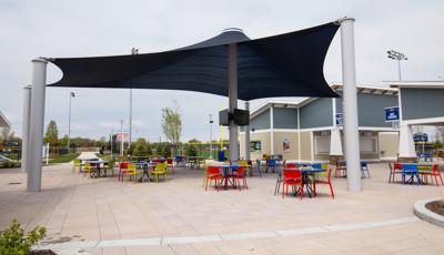 Tables and chairs of primary colors sit under a large navy blue custom shade structure with TV monitors on the center posts in the concession area of a sports center. 