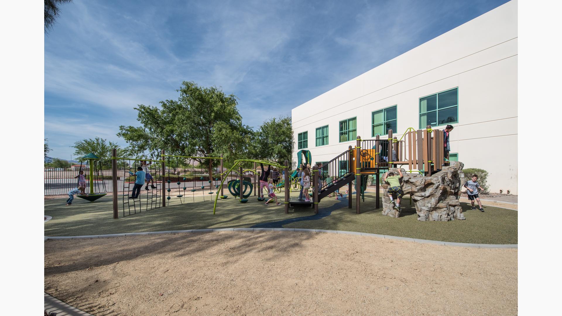 At Silver Sands Montessori Charter School playground, a boy with a cast climbs a net, while two children play on a Sol Spinner. A larger group plays on the main PlayBooster structure.