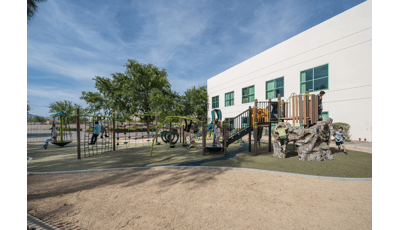 At Silver Sands Montessori Charter School playground, a boy with a cast climbs a net, while two children play on a Sol Spinner. A larger group plays on the main PlayBooster structure.