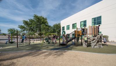 At Silver Sands Montessori Charter School playground, a boy with a cast climbs a net, while two children play on a Sol Spinner. A larger group plays on the main PlayBooster structure.