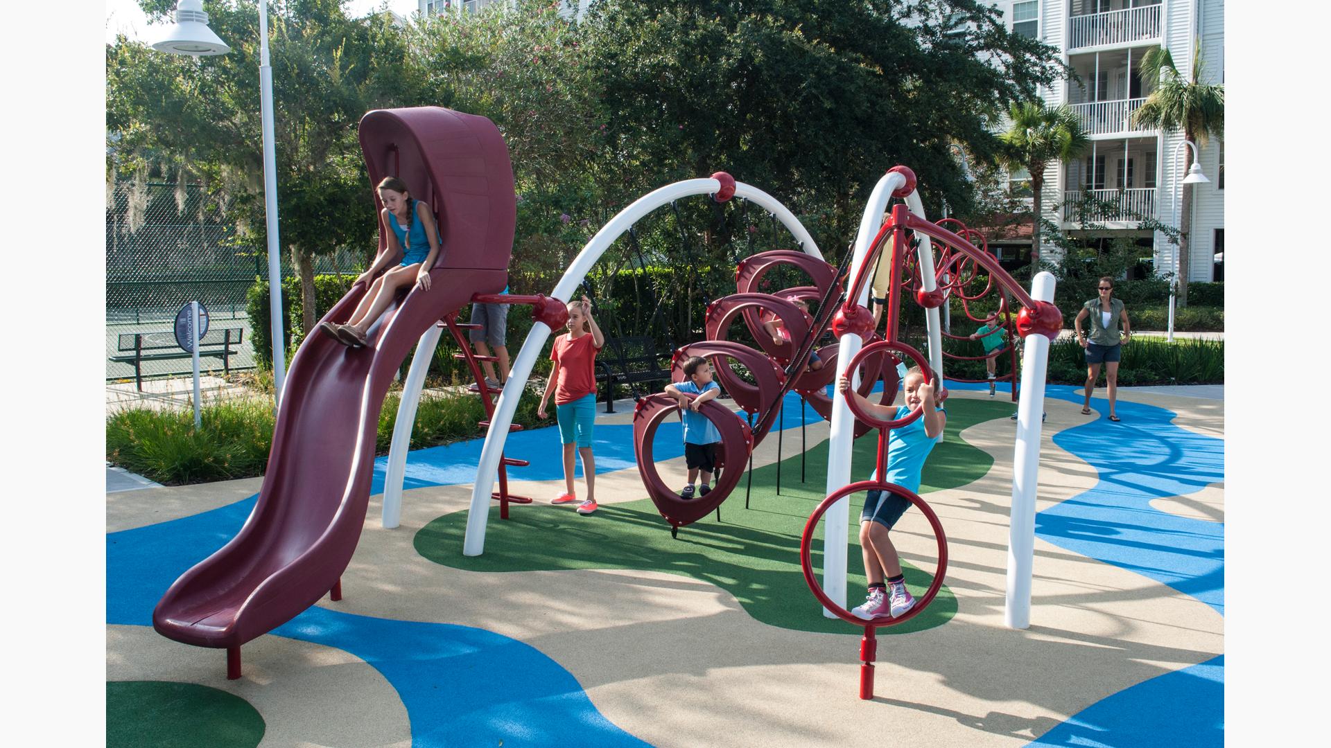 A manufactured surface on the ground. A girls rides down the Evos slide. Other kids play on the other parts of the structures as an adult looks on. 
