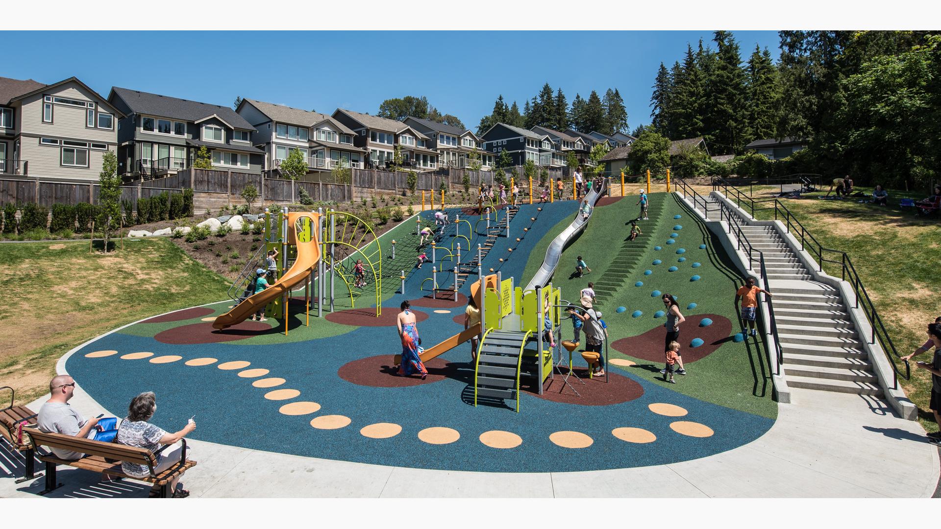 Two people on a bench sit and watch families playing on playground. Toddlers play with their parents on PlayShaper play structure. Older kids climb on PlayBooster play structure. The Nestled on the side of a hill there is a line for the metallic slide that goes that runs all the way down the hill to the bottom.