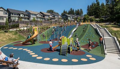 Two people on a bench sit and watch families playing on playground. Toddlers play with their parents on PlayShaper play structure. Older kids climb on PlayBooster play structure. The Nestled on the side of a hill there is a line for the metallic slide that goes that runs all the way down the hill to the bottom.