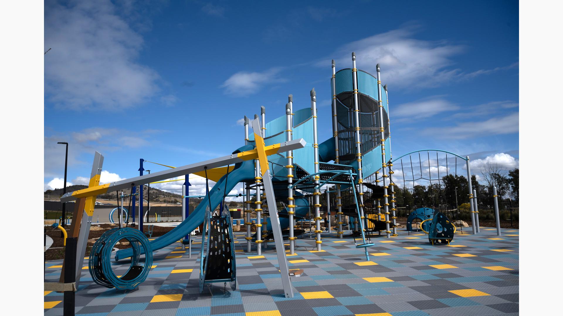 A open playground area with large tower and other surrounding play activities with the ground surfacing made of multi-colored square tiles.