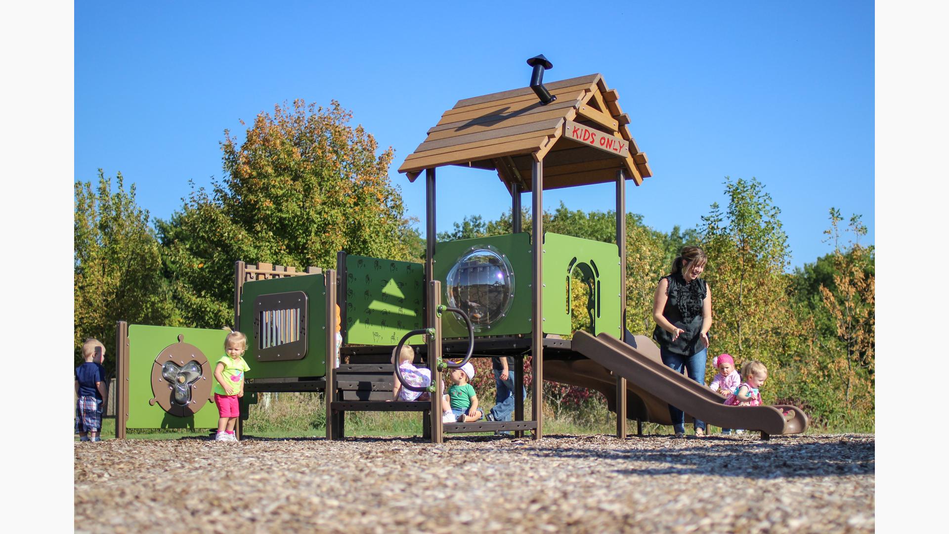Young children play on a toddler accessible playground, a mother claps for a girl riding down the structures slide. 