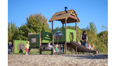 Young children play on a toddler accessible playground, a mother claps for a girl riding down the structures slide. 