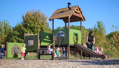 Young children play on a toddler accessible playground, a mother claps for a girl riding down the structures slide. 