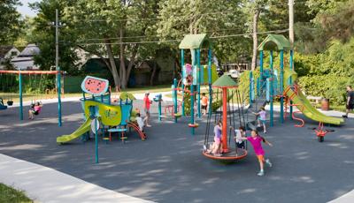 Children play at a play area with two tower structures equipped with slides and climbers for older children and a small modern design house structure for smaller children. Other children play on a swing set and multi rider spinner. 