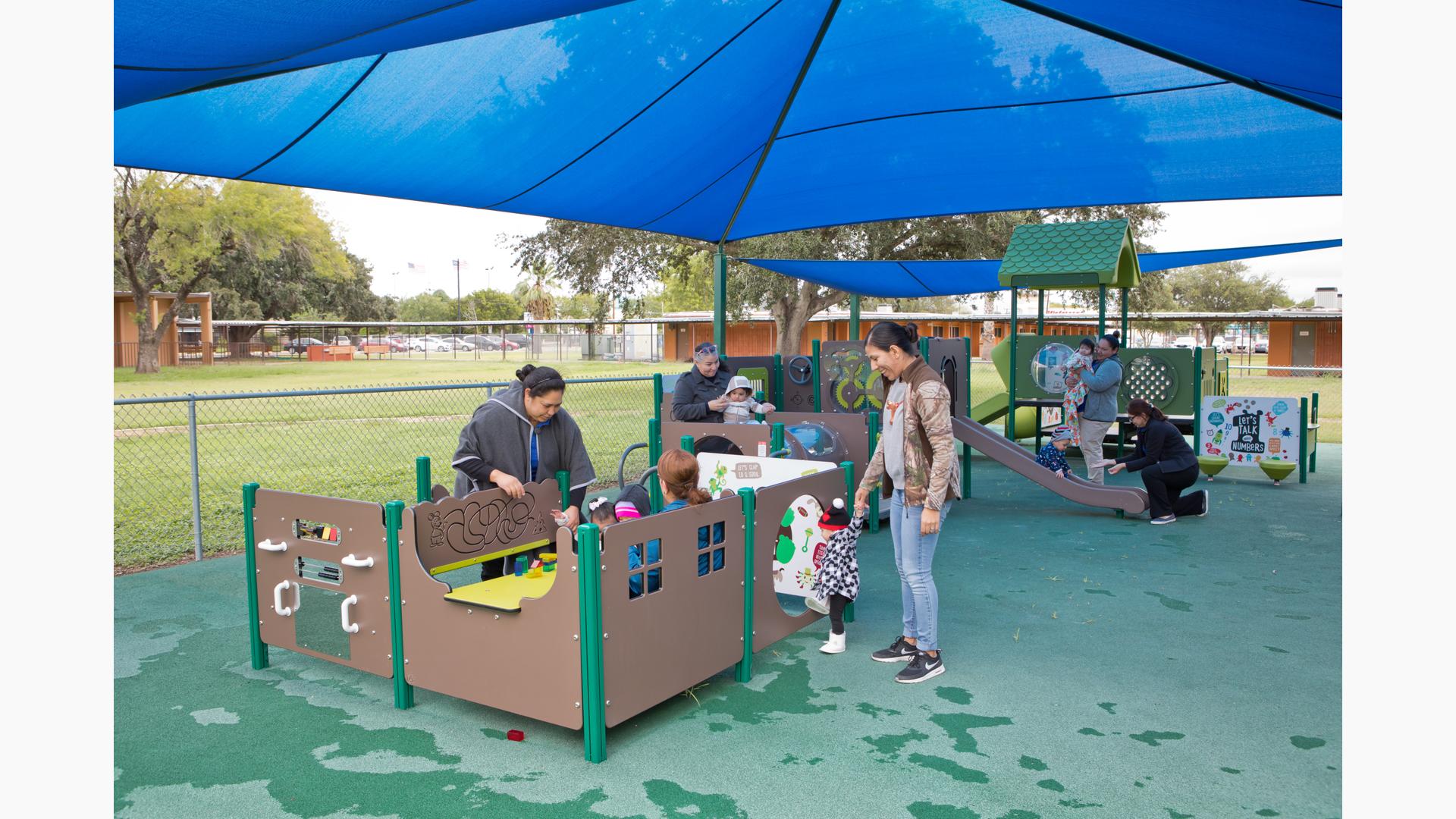 Mothers play with their young children at play panels all connecting to create a play structure. A large blue shade covers the play area.