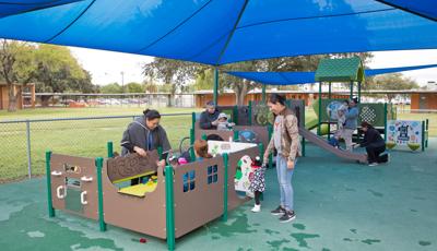 Mothers play with their young children at play panels all connecting to create a play structure. A large blue shade covers the play area.