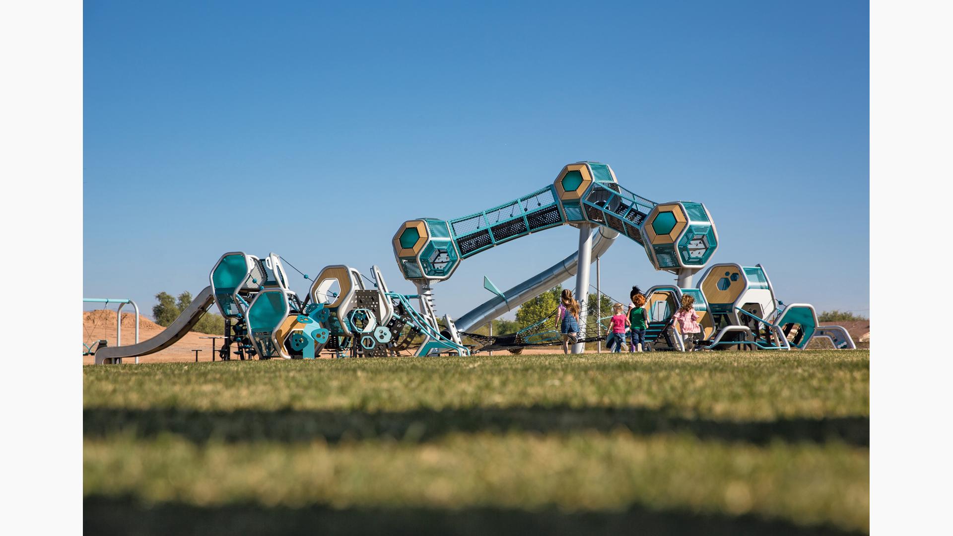 Three separate futuristic like play structures have hexagonal play pods all connected by climbers, slides, and belted pathways. One structure is three towers with a single hexagonal pod on the top, enclosed crawl tunnels connect the play pods.