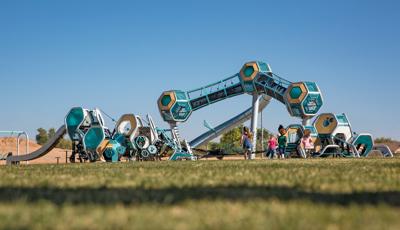 Three separate futuristic like play structures have hexagonal play pods all connected by climbers, slides, and belted pathways. One structure is three towers with a single hexagonal pod on the top, enclosed crawl tunnels connect the play pods.