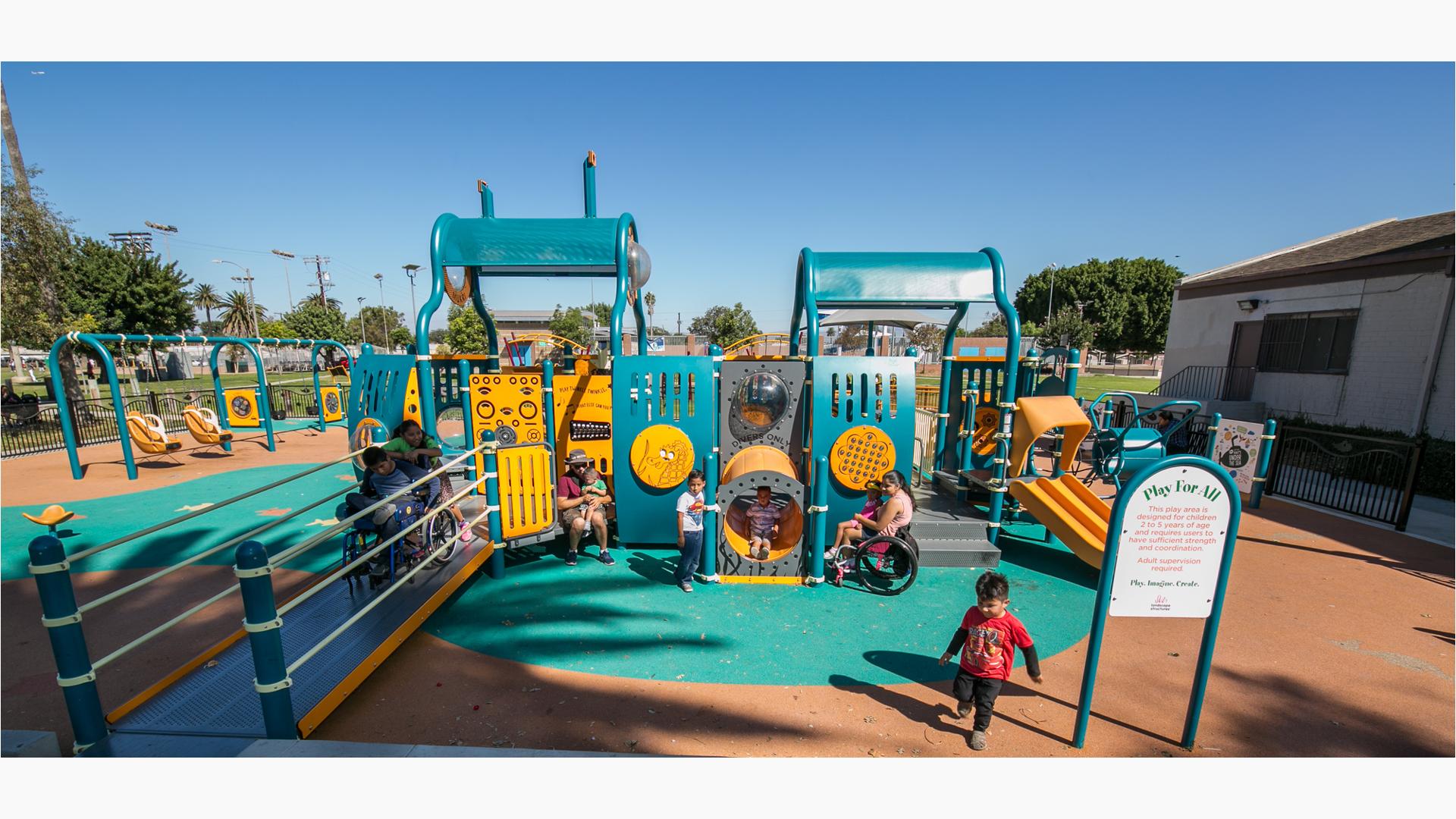 small kids run and play around a teal and orange ramped playground with surfacing. Swings and a building are in the background of a warm blue sky.