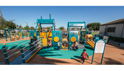 small kids run and play around a teal and orange ramped playground with surfacing. Swings and a building are in the background of a warm blue sky.
