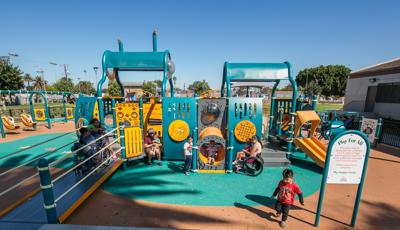 small kids run and play around a teal and orange ramped playground with surfacing. Swings and a building are in the background of a warm blue sky.