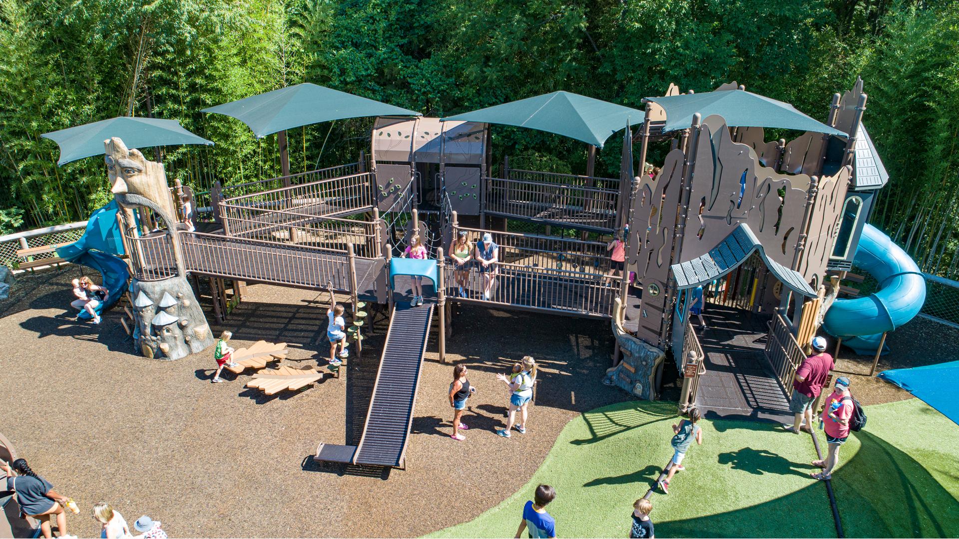 A large nature themed play structure filled with playing families. Extra wide inclusive accessible ramps connect the raised playground decking to different play experiences. The fenced in play area butts up next to a lush green woods.